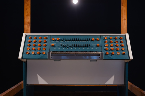 A dashboard machine, in a corner, with an old yet colorfull aspect. The machine is white and turquoise and has a lot of black and orange buttons.